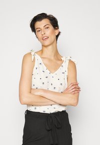 Top branco sem mangas com padrão floral azul, decote em V, e alças ajustáveis; combinado com uma saia preta de cintura alta texturizada.