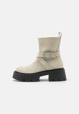 Beżowe botki za kostkę wykonane z zamszu, z szeroką, fakturowaną czarną podeszwą. Posiadają boczny pasek z klamrą oraz pętelkę do zakupu na pięcie.