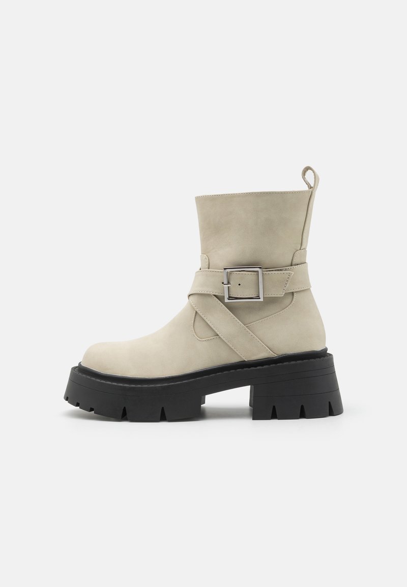 Beige ankle boot made of suede with a wide, textured black sole. Features a side buckle strap and a loop pull tab at the heel.