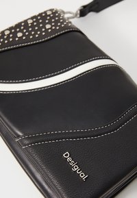 Borsa a tracolla in pelle nera con strisce bianche, borchie decorative sulla pattina, cuciture bianche a contrasto e un logo argentato.