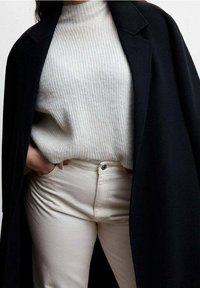 Pull en ribbed gris clair, manteau noir et jeans blanc cassé. Le pull a un col haut ; le manteau présente des revers marqués. Textures subtiles visibles.