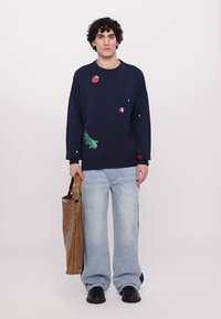 Marineblauer Pullover mit farbenfrohen gestickten Akzenten, kombiniert mit hellblauen Jeans im weit geschnittenen Stil. Das Model hält eine braune Tragetasche. Schwarze Schuhe.