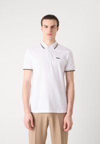 Hvit polo skjorte med svarte og beige detaljer på kragen og ermene, med tre knapper og en liten "BOSS" logo på brystet.