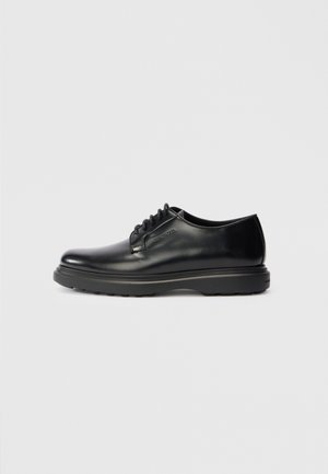 Zapato oxford de piel negra con cordones y suela gruesa, con "WATERMAN" grabado en el lateral, mostrado de perfil sobre un fondo gris claro.