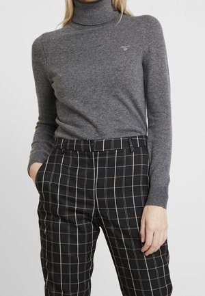 Vrouw die een nauwsluitende grijze coltrui en zwarte broek met witte en bruine ruitjes draagt, staat met één hand in haar zak.
