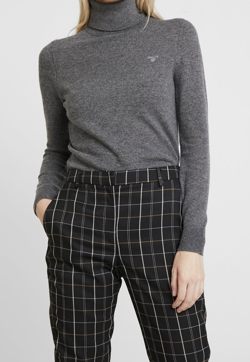 Femme portant un pull col roulé gris ajusté et un pantalon noir à carreaux blancs et marron, debout avec une main dans la poche.