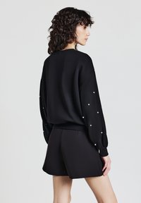 Maglione nero a maniche lunghe con dettagli di perle sulle maniche, abbinato a pantaloncini neri. Il tessuto sembra liscio e leggero.