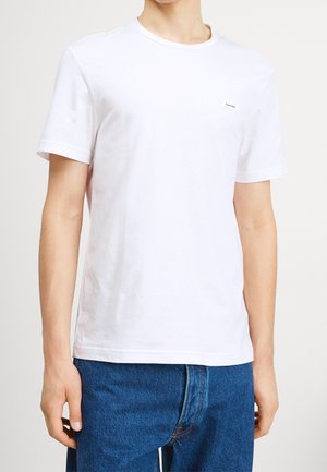 Biały bawełniany t-shirt z krótkimi rękawami, okrągłym dekoltem i małym logo na lewej piersi. W zestawie z niebieskimi jeansami.