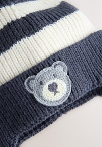 Gestrickte Mütze mit marineblauen und cremefarbenen Streifen. Sie verfügt über einen strukturierten Stoff und einen braun geformten, grauen und weißen bestickten Bären-Aufnäher am Bündchen.