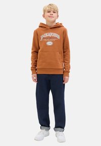 Brown hooded sweatshirt with front pocket and white logo text, paired with dark blue relaxed-fit pants and white sneakers.