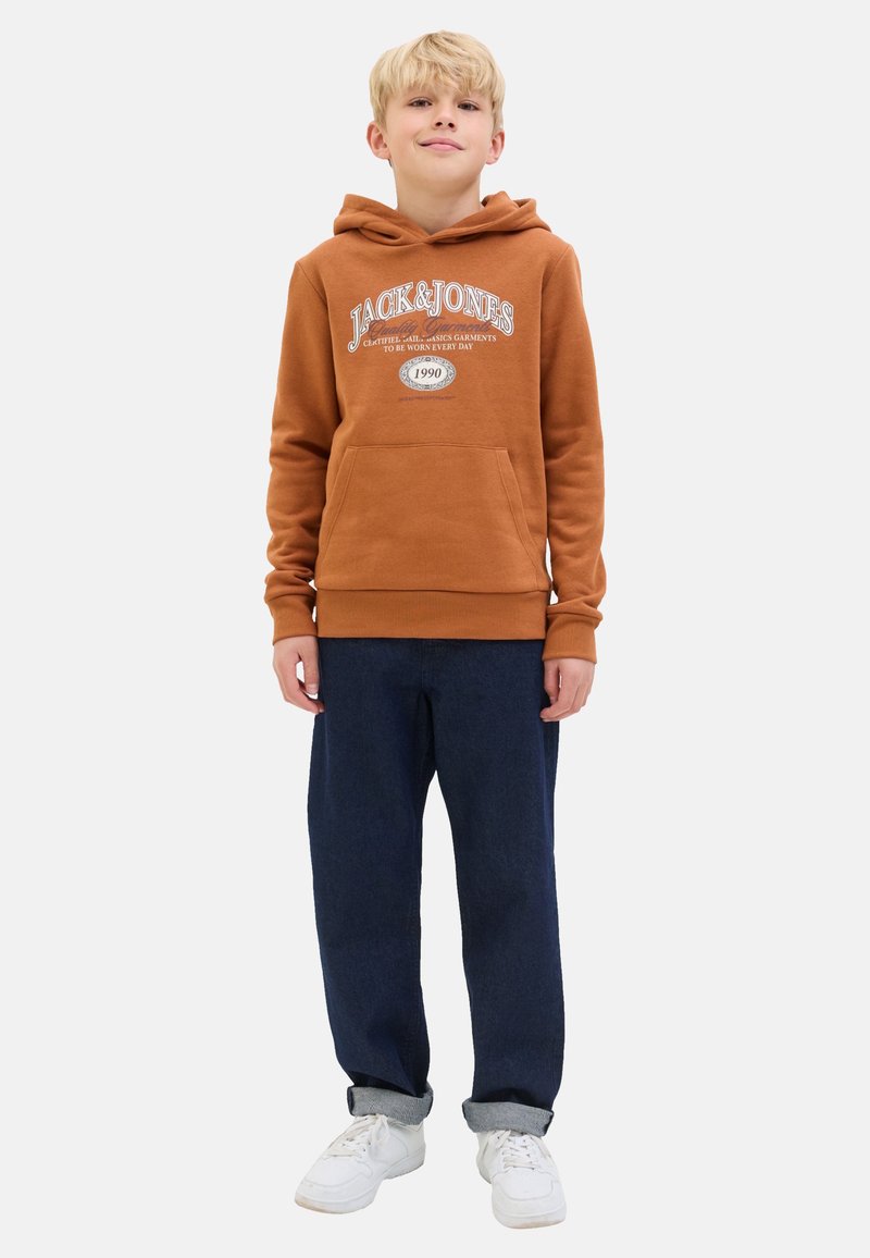 Brown hooded sweatshirt with front pocket and white logo text, paired with dark blue relaxed-fit pants and white sneakers.