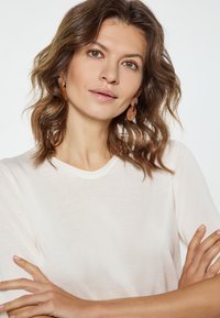 Creme, ribbet strikket top med rund halsudskæring. Modellen bærer gennemsigtige, runde øreringe. Håret er bølget og rammer ansigtet. Blødt lys.