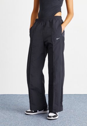 Personne portant un pantalon noir ample Nike avec une taille élastique et des baskets noires et blanches, debout avec les mains dans les poches.