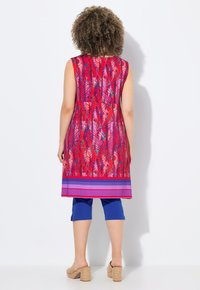 Jasný, vzorovaný bezrukávový tunik s odstíny červené, fialové a modré. V kombinaci s modrými capri kalhotami a béžovými klínovými botami.