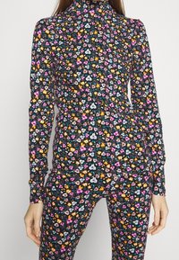 Frau trägt einen enganliegenden schwarzen Rollkragenpullover und eine Hose mit einem lebhaften kleinen Blumenmuster in pink, orange, weiß und blautönen.