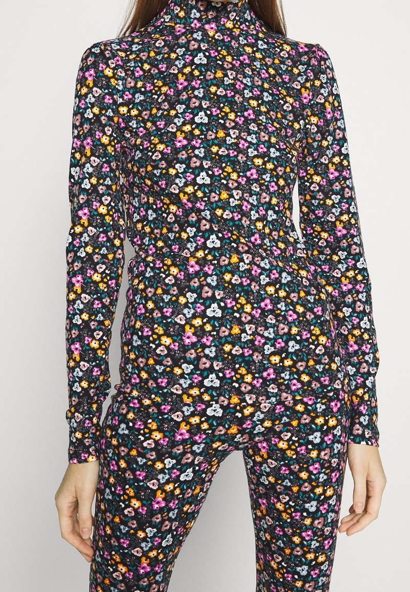 Frau trägt einen enganliegenden schwarzen Rollkragenpullover und eine Hose mit einem lebhaften kleinen Blumenmuster in pink, orange, weiß und blautönen.