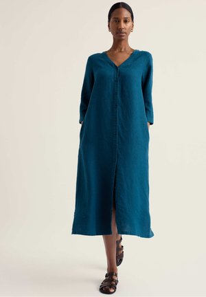 Femeie purtând o rochie lungă teal cu nasturi, mâneci trei sferturi și sandale negre cu barete, stând în fața unui fundal simplu.