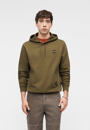 Junger Mann trägt einen olivgrünen Hoodie mit einer Fronttasche und einem Logo auf der Brust, kombiniert mit grauen Jeans und einem silbernen Armband.