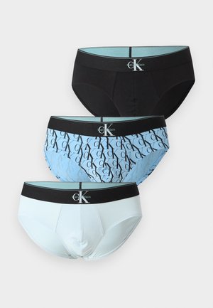 Trzy męskie slipy w kolorze czarnym, jasno niebieskim z czarnym wzorem "ck" oraz jednolite białe, każdy z czarną gumą w pasie z białym logo Calvin Klein.