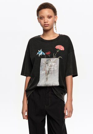 Joven con una camiseta negra holgada con un diseño de flores dentro de una bolsa translúcida y pantalones negros, de pie frente a un fondo blanco.