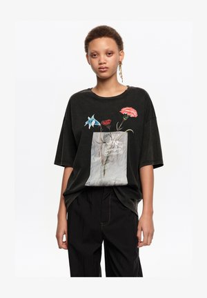 Jeune personne portant un t-shirt noir ample avec un motif de fleurs dans un sac translucide et un pantalon noir, debout devant un fond blanc.