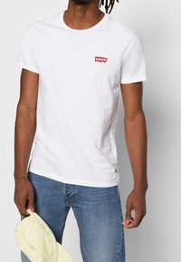 Bílá bavlněná tričko s červeným logem Levi's na levém prsu, v kombinaci s modrými džíny a držící světlou žlutou čepici.