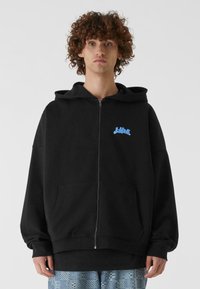 Sudadera con cremallera negra, con un bolsillo delantero y un logo azul claro en el pecho; presenta un diseño espacioso y una textura de tela suave.