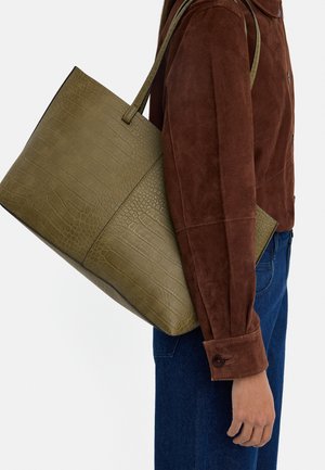 Osoba w brązowej zamszowej kurtce i niebieskich dżinsach niosąca na ramieniu dużą oliwkowozieloną torbę na ramię z wzorem krokodyla.