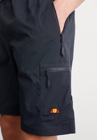 Marineblå shorts laget av lett materiale, med en glidelåslomme, snor detaljer og et fargerikt logo-patch på siden.