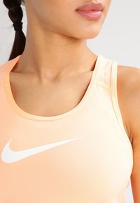Różowo-brzoskwiniowy sportowy tank top wykonany z gładkiego, elastycznego materiału. Posiada okrągły dekolt, cienkie ramiączka oraz biały logo Nike z przodu.