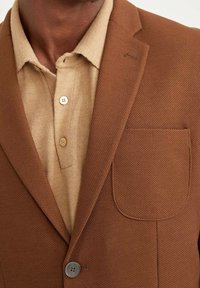 Bruine blazer met textuur en een enkele knoopsluiting. Beige gebreide poloshirt eronder met twee knopen en een kraag. Voorzak zichtbaar.