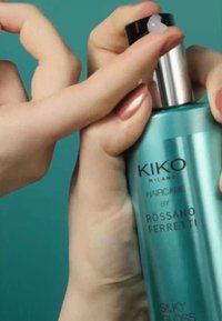 Produit de soins capillaires KIKO Milano dans une bouteille turquoise avec un capuchon argenté, étant pulvérisé par un embout vaporisateur. L'étiquette indique "Brillance Soyeuse."