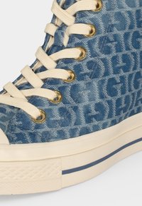 Baskets montantes en denim avec lacets blancs, œillets dorés, motif texte "GIGI" bleu foncé répété et semelle en caoutchouc blanc cassé avec bande bleue.