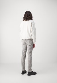 Jasnokremowa bluza, szare jeansy o fakturze oraz czarne obuwie. Strój charakteryzuje się luźnym krojem, widocznymi przeszyciami oraz subtelnym efektem postarzenia.