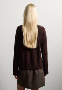Pull en tricot marron avec un ourlet et des manches côtelés, associé à une jupe marron texturée, vu de dos contre un mur uni.