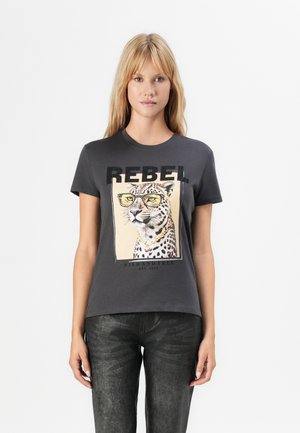 Femme blonde portant un T-shirt gris foncé avec un imprimé léopard, des lunettes et une chaîne dorée, un pantalon noir texturé, se tenant devant un fond blanc.