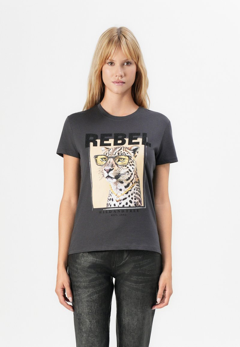 Blonde woman wearing dark gray T-shirt with leopard print wearing glasses and a gold chain, black textured pants, standing against white background.