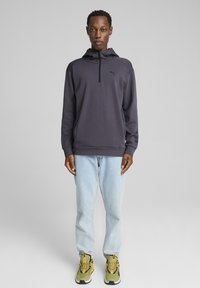 Sudadera de felpa gris oscuro para hombre con media cremallera, logotipo negro, combinada con jeans rectos de color azul claro y zapatillas con detalles amarillos.