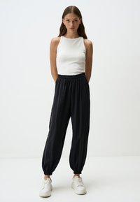 Pantaloni neri a vestibilità ampia con vita elastica e polsini, abbinati a una canotta bianca senza maniche. Modello che indossa sneaker bianche. Sfondo minimale.