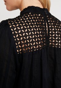 Schwarze Bluse mit aufwendiger Spitzenverzierung auf der Rückseite in kreisförmigen Mustern. Der Rüschenausschnitt und der Knopfverschluss heben die Designmerkmale hervor.