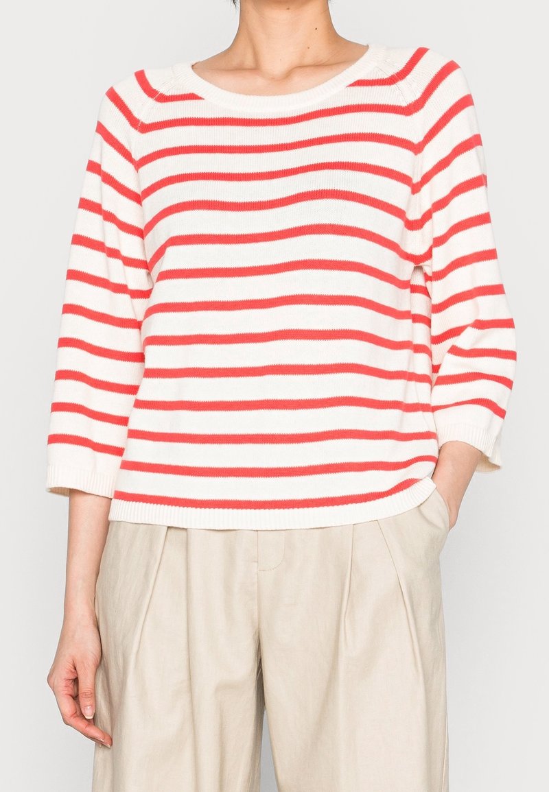 Personne portant un pull en maille blanc à rayures horizontales rouges et un pantalon plissé taille haute beige, une main dans la poche.