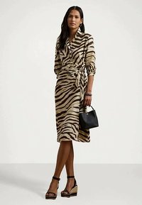 Γυναίκα με φόρεμα περιτυλίγματος με σχέδιο ζέβρας, μαύρες πλατφόρμες και κρατώντας μια μικρή μαύρη τσάντα, σε φόντο απλό.