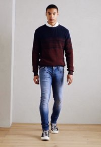 Suéter a rayas en rojo oscuro y azul marino, jeans azules, zapatillas negras. El modelo está de pie sobre un suelo de madera con un fondo de pared gris claro.