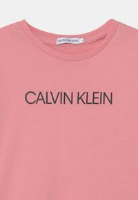 Camiseta de algodón rosa con cuello redondo y el texto "CALVIN KLEIN" impreso en negro. Textura suave, diseño casual.