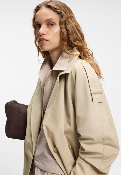 Jeune femme aux cheveux blonds ondulés portant un trench-coat beige et une chemise claire, tenant une pochette marron foncé sur son épaule.