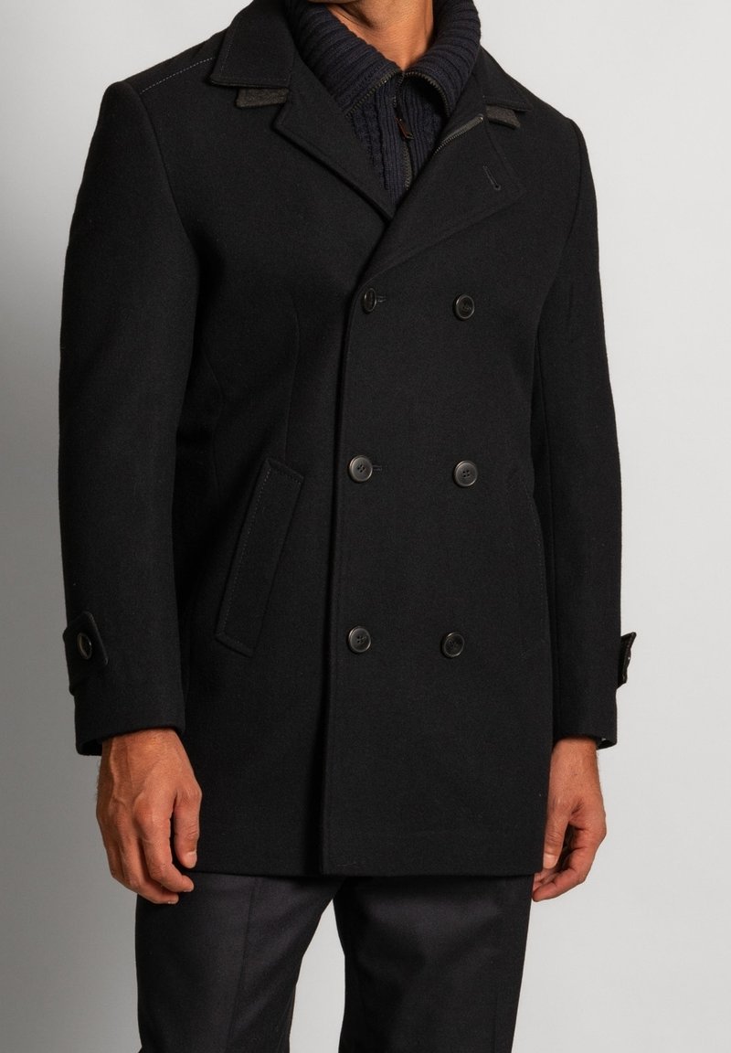 Homme portant un manteau caban noir à double boutonnage sur un pull en tricot noir zippé, debout devant un fond uni.