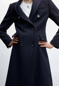 Cappotto doppiopetto in lana navy con colletto a punta, due tasche frontali e grandi bottoni neri. Presenta una silhouette aderente e una texture elegante.