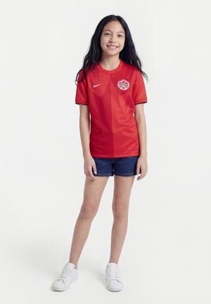 Pige står og smiler, iført en rød Canada fodboldtrøje, mørkeblå shorts og hvide sneakers mod en ensartet hvid baggrund.