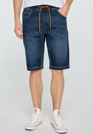 Mężczyzna noszący ciemnoniebieskie szorty jeansowe z pomarańczowym ściągaczem i białe sneakersy za kostkę na jednolitym tle.