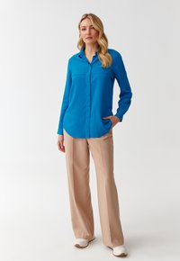 Blue long-sleeve shirt with a collar and two chest pockets, paired with light beige wide-leg trousers and white sneakers.